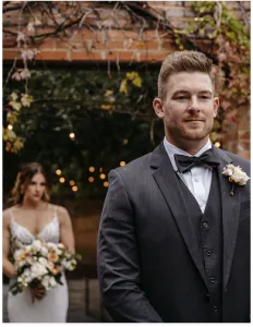 Groom in a suit standing outdoors at a wedding venue with the bride in the background.