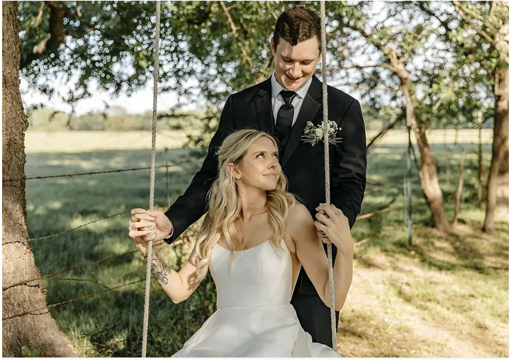 Bride sitting on a tree swing while her groom stands behind her during an outdoor wedding portrait.
