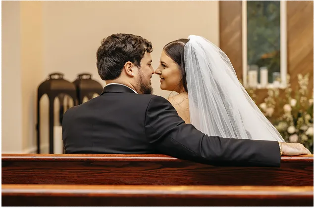 Newlywed bride and groom sitting together in a church pew and looking at each other after their ceremony.